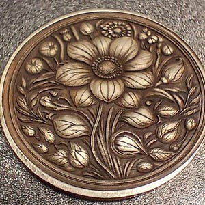 May include: A silver-toned metal coin featuring an intricate design of a flower with leaves and buds. The flower is positioned in the centre of the coin and is surrounded by leaves and buds.