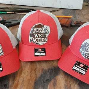 Op de afbeelding: Drie rode truckerpetten met witte mesh-achterkanten. Elke pet heeft een andere patch: "I'm Just a Forester", "My Pole Needs Some Wet Action" en "Morningwood Camp Grounds". Elke pet heeft een Richardson 115-label.