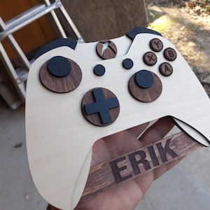 Op de afbeelding: Een houten Xbox-controller wanddecoratie met een bruine houten basis en zwarte knoppen. De naam "ERIK" is gegraveerd op de basis.