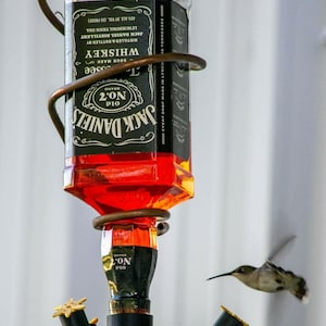 May include: A repurposed Jack Daniel's whiskey bottle hummingbird feeder. The feeder is made from a black and white bottle with a copper-colored wire frame. A hummingbird is flying near the feeder.