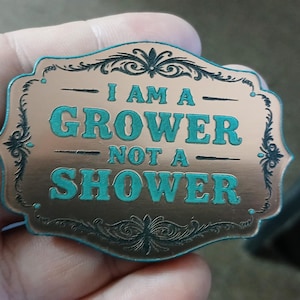 May include: A copper-colored metal badge with the phrase "I AM A GROWER NOT A SHOWER" in teal lettering. The badge has a decorative border with floral and scroll designs. The badge is held in a hand.