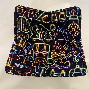 May include: A black pot holder with a neon-colored camping themed pattern. The pattern includes a campfire, trees, a compass, a tent, and hiking boots.