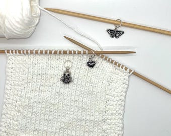 May include: White knitting project with three stitch markers. The markers are a silver skull, a silver heart, and a silver butterfly.