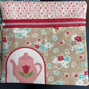May include: A brown fabric zippered pouch with a red zipper and a floral pattern. The pouch features a red and white teapot with a red tulip design.