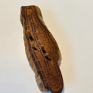 May include: A handcrafted cribbage board made from a piece of natural wood. The board features a series of drilled holes for scoring and includes wooden pegs. The wood grain is visible, showcasing a mix of light and dark brown tones.