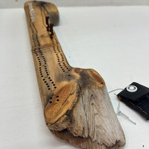 May include: A wooden cribbage board crafted from driftwood with a natural finish. The board features a traditional design with holes for pegs and a scoring track. The board comes with a small black leather pouch with a white button.