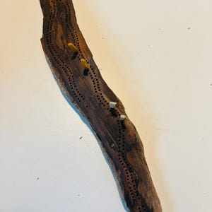 May include: A handcrafted cribbage board made from driftwood. The board is dark brown with a natural wood grain and features rows of drilled holes for pegs. Three pegs are inserted: two yellow and one silver.