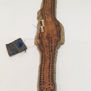 May include: A wooden cribbage board with a natural wood finish. The board is shaped like a long, narrow rectangle with rounded edges. There are holes drilled into the board for pegs. The board is accompanied by a small brown leather pouch with a blue button.
