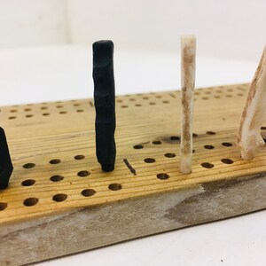 May include: A wooden cribbage board with four pegs inserted. Two black pegs and two off-white pegs are in the board. The board is made of light-coloured wood with evenly spaced holes for the pegs.