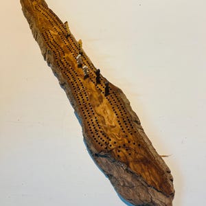 Op de afbeelding: Een handgemaakt cribbagebord van drijfhout, met een natuurlijke bruine tint. Het heeft talrijke geboorde gaten en wordt geleverd met kleine, gekleurde pinnen. Dit bord is ontworpen voor het traditionele kaartspel en biedt een rustieke uitstraling.