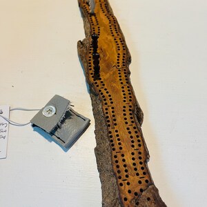 May include: A cribbage board made from a piece of natural wood, with drilled holes for pegs. It is accompanied by a small grey leather pouch with a button fastening. The board has a rustic, organic appearance.