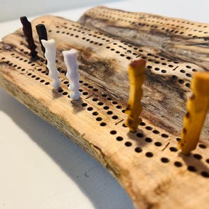 May include: A wooden cribbage board with a live edge, showing a natural, rustic aesthetic. The board is drilled with holes for pegs, and several pegs in brown, white, and yellow are inserted. The board's dimensions are approximately 30 cm in length.