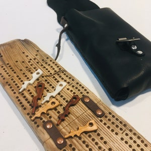 May include: A rustic cribbage board made of wood, with numerous holes and a selection of wooden pegs. A black leather pouch is visible, likely for storing the pegs, suggesting a complete game set.