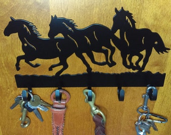 May include: Black metal wall mounted key holder with a silhouette of three horses running. The key holder has five hooks and is hanging on a wooden wall.