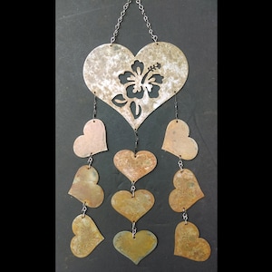 May include: A metal heart-shaped wind chime with a cutout hibiscus flower design. The chime has seven smaller hearts hanging from it, each with a different shade of brown.
