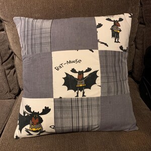 May include: A square throw pillow featuring a patchwork design of alternating grey plaid fabric squares and white fabric squares printed with a cartoon moose dressed as a superhero. The text "Bat-Moose" is printed above the central moose figure.