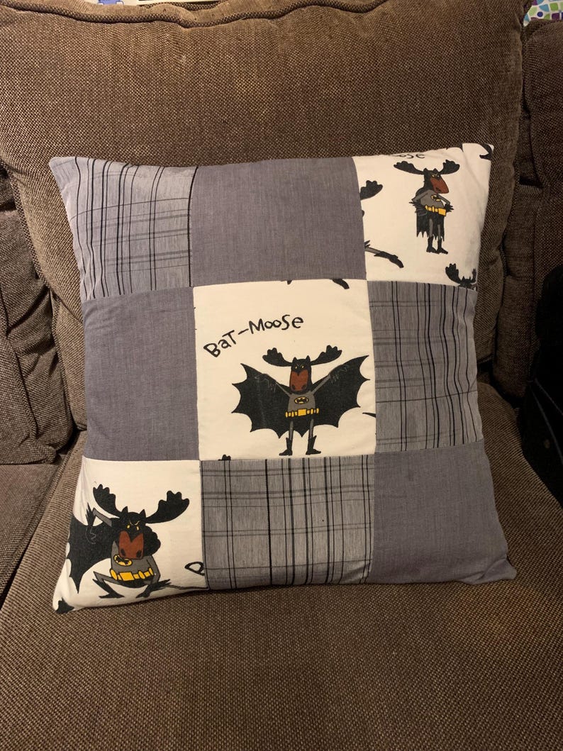 May include: A square throw pillow featuring a patchwork design of alternating grey plaid fabric squares and white fabric squares printed with a cartoon moose dressed as a superhero. The text "Bat-Moose" is printed above the central moose figure.