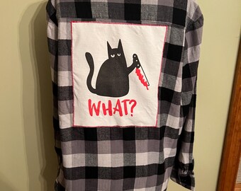 Puede incluir: Una camisa de manga larga con botones y un patrón de cuadros en negro, gris y blanco. En la parte posterior hay un parche cuadrado blanco cosido con la ilustración de un gato negro sosteniendo un cuchillo con manchas de sangre roja y el texto rojo WHAT?