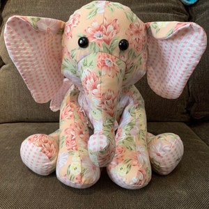 May include: A pink and floral patterned stuffed elephant toy with black button eyes. The elephant is sitting on a brown couch.