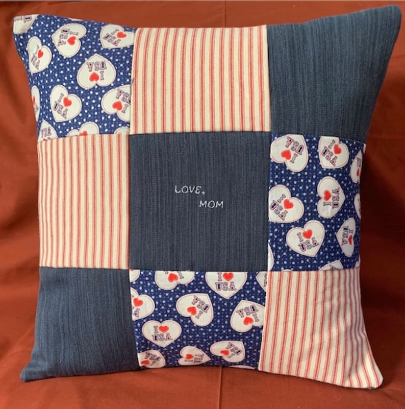 May include: A square patchwork pillow featuring a grid of nine fabric squares. The design includes blue fabric with white polka dots and heart-shaped 'I Love USA' prints, red and white striped fabric, and solid blue fabric with 'LOVE, MOM' embroidered.