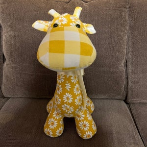May include: A yellow and white stuffed giraffe with a daisy pattern and a checkered pattern on the head. The giraffe is sitting on a brown couch.