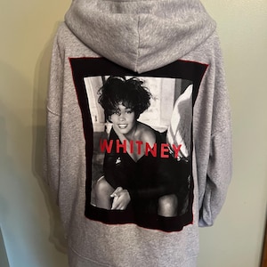 May include: A gray hoodie with a black and white image of Whitney Houston on the back. The image is framed with a black border and red trim, with the name "WHITNEY" in red letters.