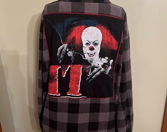 Puede incluir: Una camisa de manga larga con botones y estampado de cuadros grises y negros. El panel trasero muestra un gran estampado gráfico de un payaso con pelo rojo brillante, cara blanca y dedos largos, sobre el texto rojo IT.
