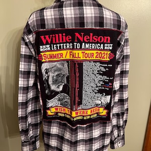 May include: A black and white plaid flannel shirt with a large black patch on the back. The patch features the text "Willie Nelson" and "Letters to America" with a tour schedule and a portrait of Willie Nelson.