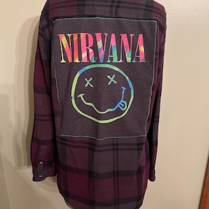 May include: A burgundy and black plaid flannel shirt with a gray patch on the back. The patch features the word "NIRVANA" in rainbow colors above a smiley face with crossed-out eyes and a tongue sticking out.