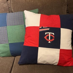 May include: Two square throw pillows made of patchwork fabric. The front pillow features a grid of red, white, and navy blue squares, with a Minnesota Twins logo and text printed on the center navy square. The back pillow has blue and green squares.