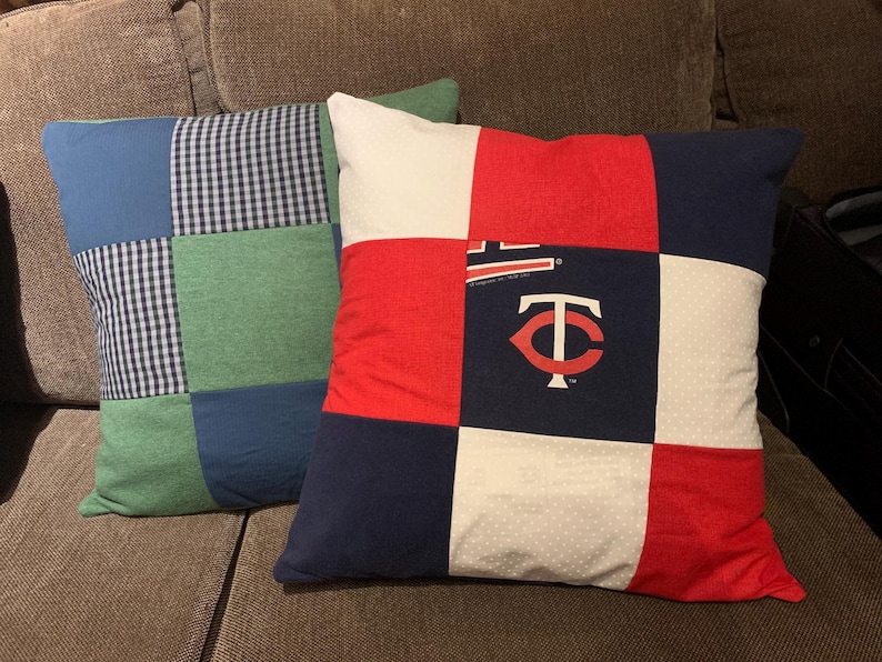 May include: Two square throw pillows made of patchwork fabric. The front pillow features a grid of red, white, and navy blue squares, with a Minnesota Twins logo and text printed on the center navy square. The back pillow has blue and green squares.