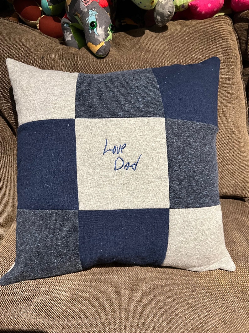 May include: A square patchwork pillow constructed from nine fabric squares in alternating shades of navy blue, dark heather grey, and light grey. The center square features the embroidered text Love Dad in dark blue thread on a light grey background.