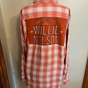 May include: Orange and white plaid button-down shirt with a brown patch on the back. The patch features the text "The Legend Lives On" above "Willie Nelson" in a vintage font. Two stars flank the name.