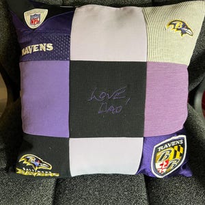 May include: A square patchwork pillow constructed from Baltimore Ravens sports jerseys and fabric panels in purple, black, and light gray. The center black panel features the embroidered text "Love, Dad" in purple thread, with team logos and NFL branding.