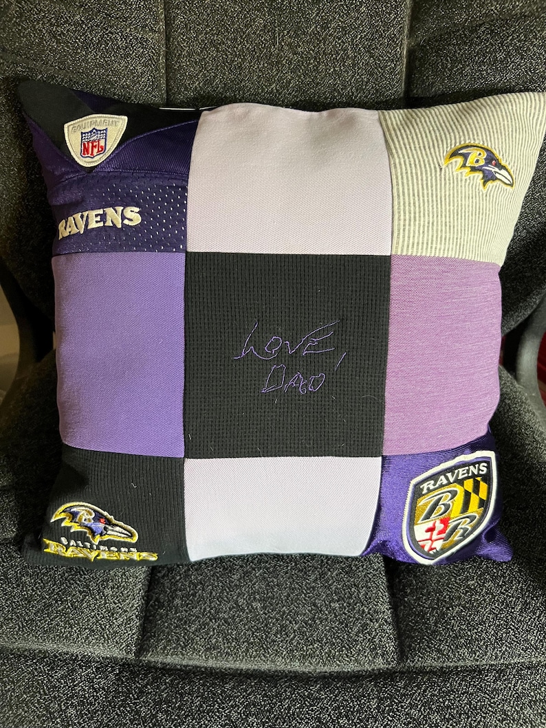 May include: A square patchwork pillow constructed from Baltimore Ravens sports jerseys and fabric panels in purple, black, and light gray. The center black panel features the embroidered text "Love, Dad" in purple thread, with team logos and NFL branding.