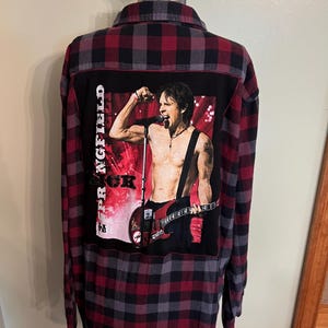 May include: Red, black, and gray plaid flannel shirt with a graphic on the back featuring a shirtless man playing a guitar and the text "Rick Springfield". The shirt has long sleeves and a button-down front.