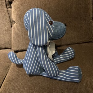 Puede incluir: Un juguete de peluche con forma de perro, de rayas azules y blancas, sentado en un sof&aacute; marr&oacute;n. El perro tiene una cinta blanca alrededor del cuello.