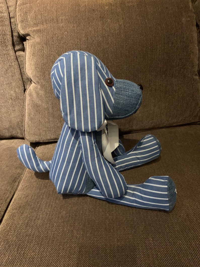 Puede incluir: Un juguete de peluche con forma de perro, de rayas azules y blancas, sentado en un sof&aacute; marr&oacute;n. El perro tiene una cinta blanca alrededor del cuello.