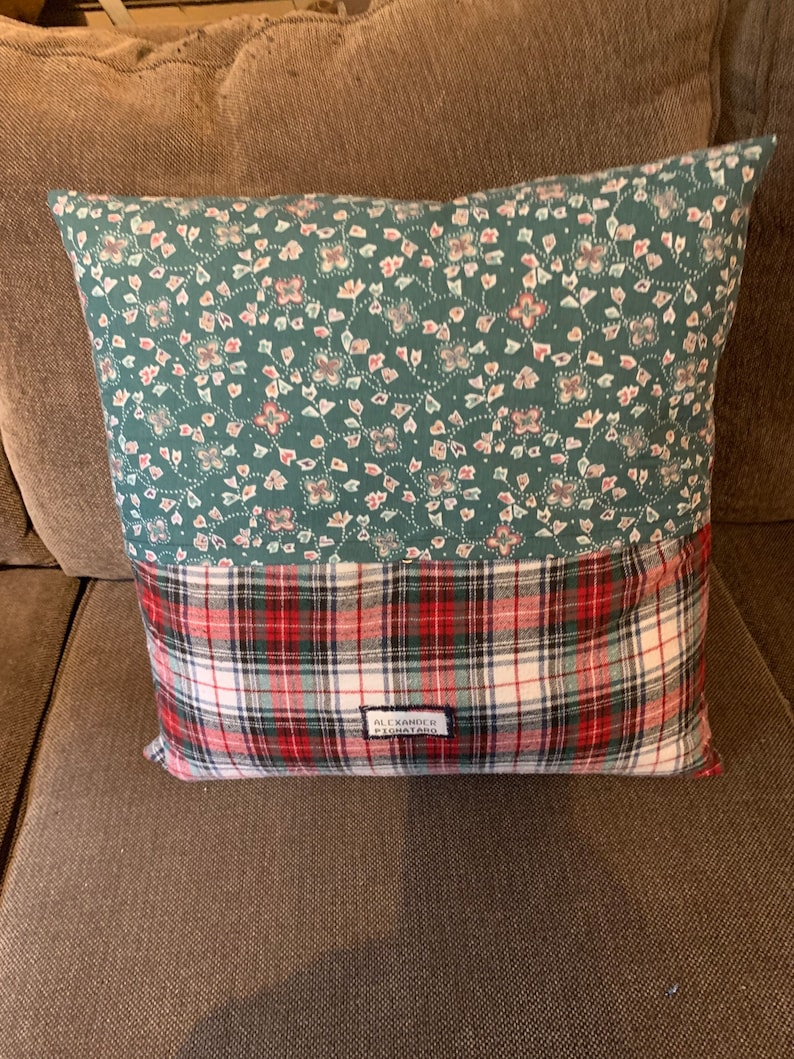 May include: A square throw pillow featuring a two-tone design. The top half is a teal fabric with a small white and pink floral pattern. The bottom half is a red, white, black, and green plaid fabric with a small rectangular label reading ALEXANDER PIGNATARO.