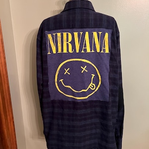 May include: A long-sleeve button-down shirt featuring a dark blue and black plaid pattern. The back panel displays a large, rectangular patch in a faded blue fabric with the yellow text NIRVANA and a yellow smiley face graphic with X eyes.