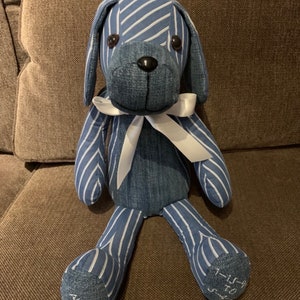 Puede incluir: Un juguete de perro de mezclilla azul y blanco a rayas con un lazo blanco. El juguete tiene ojos negros y una nariz negra. Las palabras "Alex" y "1-15-04" est&aacute;n bordadas en los pies.