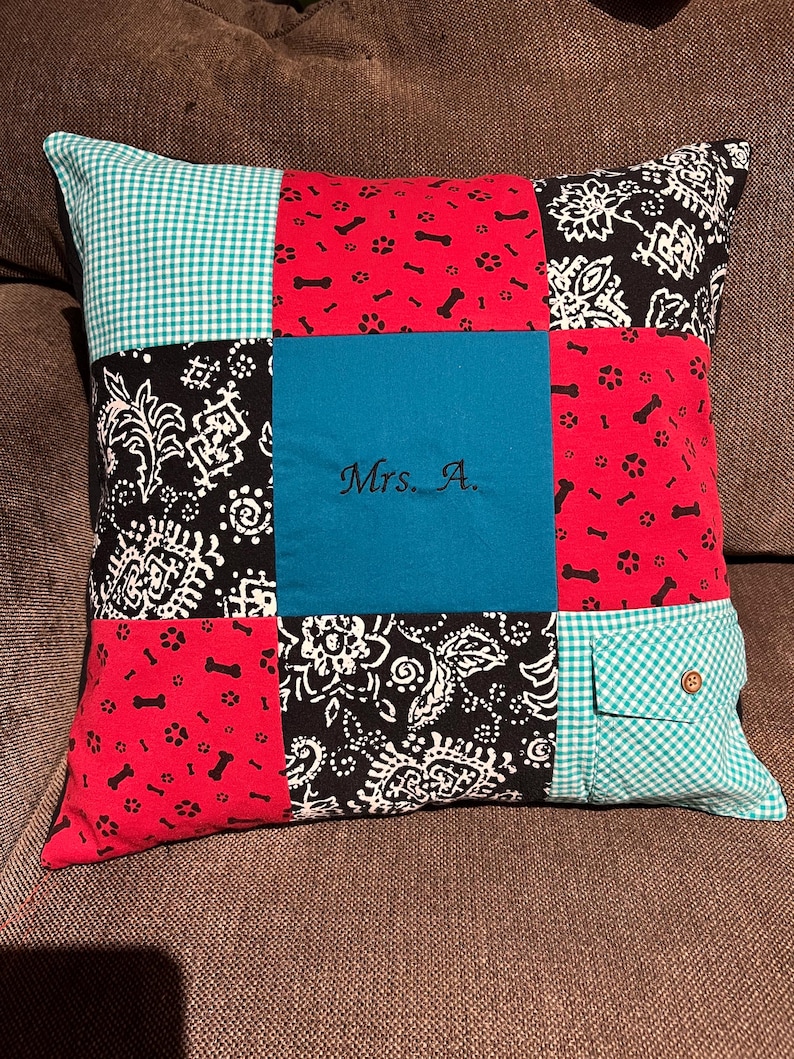 May include: A square patchwork pillow featuring nine distinct fabric panels. The design includes red fabric with black bone and paw prints, black fabric with white floral patterns, teal fabric with the embroidered text Mrs. A., and teal gingham.