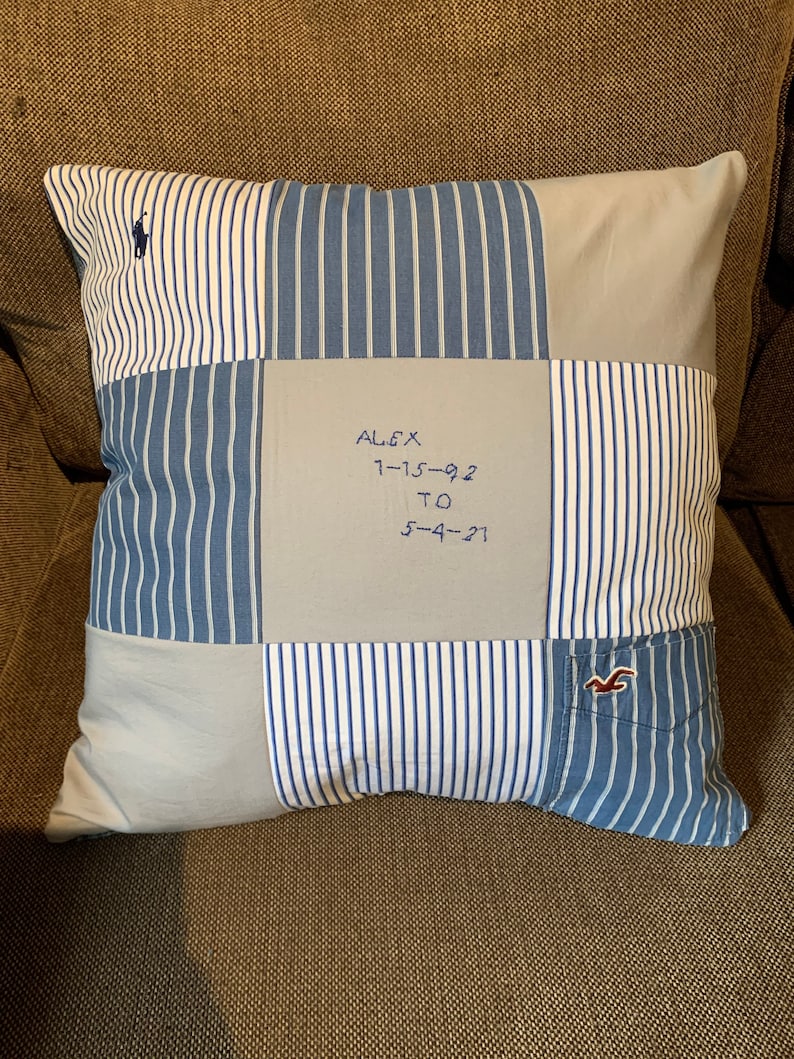 May include: A square patchwork pillow constructed from various blue and white striped and solid light blue fabric panels. The center panel features embroidered text reading ALEX 1-15-92 TO 5-4-21. A small navy blue logo is visible in the top left corner.