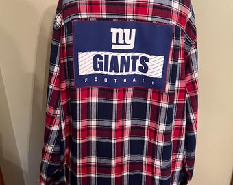 Puede incluir: Una camisa de franela de manga larga con botones en un patrón de cuadros rojos, azul marino y blancos. La parte posterior presenta un gran parche rectangular con el texto "ny GIANTS FOOTBALL" en letras blancas y azul marino sobre un fondo azul.