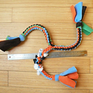 May include: A multi-coloured braided rope dog toy with two knotted ends. The rope is made of black, orange, green, and blue fabric. The toy is approximately 76 cm long.