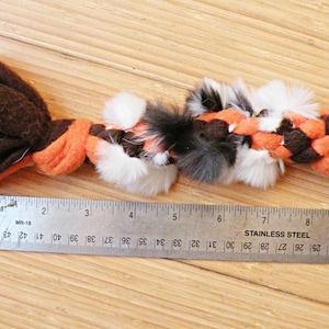 May include: A brown and orange braided dog toy with white and black fur accents. The toy is approximately 18 cm long and is measured with a ruler.