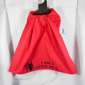 Puede incluir: Una capa de disfraz roja brillante con cuello con cordón, colgada de una percha negra. La capa presenta un gráfico negro de un diablo con un tridente y el texto "I AM A LITTLE DEVIL".