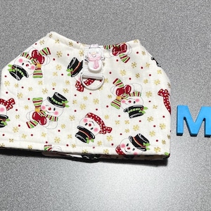 May include: A white pet harness with a repeating pattern of snowmen wearing hats and scarves. The snowmen have red and green accents. A blue letter "M" is next to the harness.