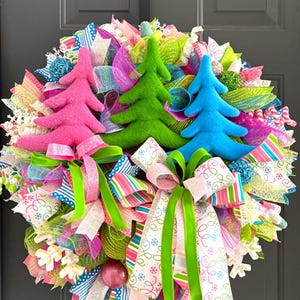 May include: A decorative wreath featuring three plush Christmas trees in bright pink, lime green, and blue. The base is composed of multi-colored mesh ribbons in pink, blue, and green, accented with white snowflake patterns and lime green bows.