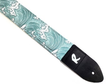 Peut inclure: Une sangle de guitare avec un fond blanc et un motif de vagues bleues. La sangle a une extrémité en cuir noir avec un logo "R" blanc.